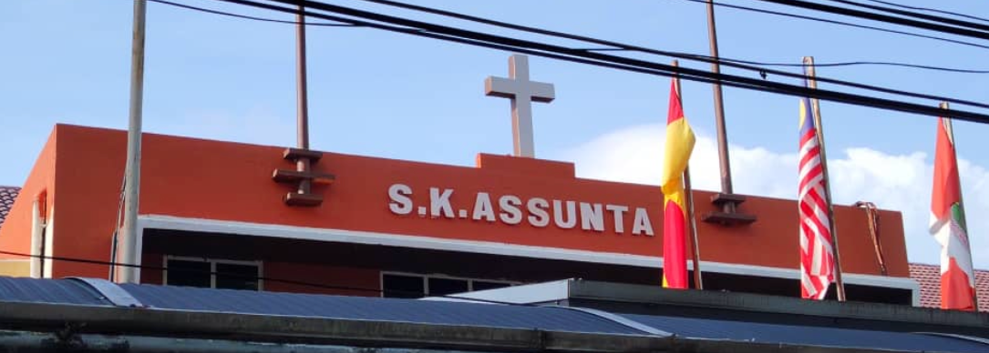 The front of SK Assunta school, with its name above the entrance, a cross on the roof and flags flying.